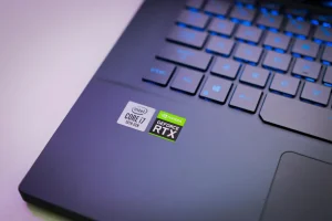 Czarny laptop gamingowy z podświetlaną klawiaturą, wyświetlający intensywną scenę z gry akcji, stoi na biurku obok myszki i słuchawek.